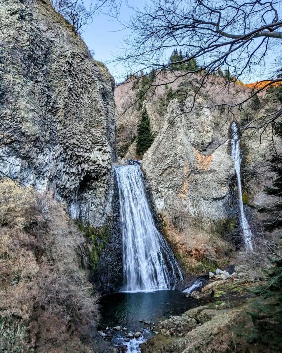 Vue sur la double cascade du Ray Pic en hiver avec les colonnes basaltiques et un lever de soleil sur les hauteurs derrnière la forêt de résineux (photo)