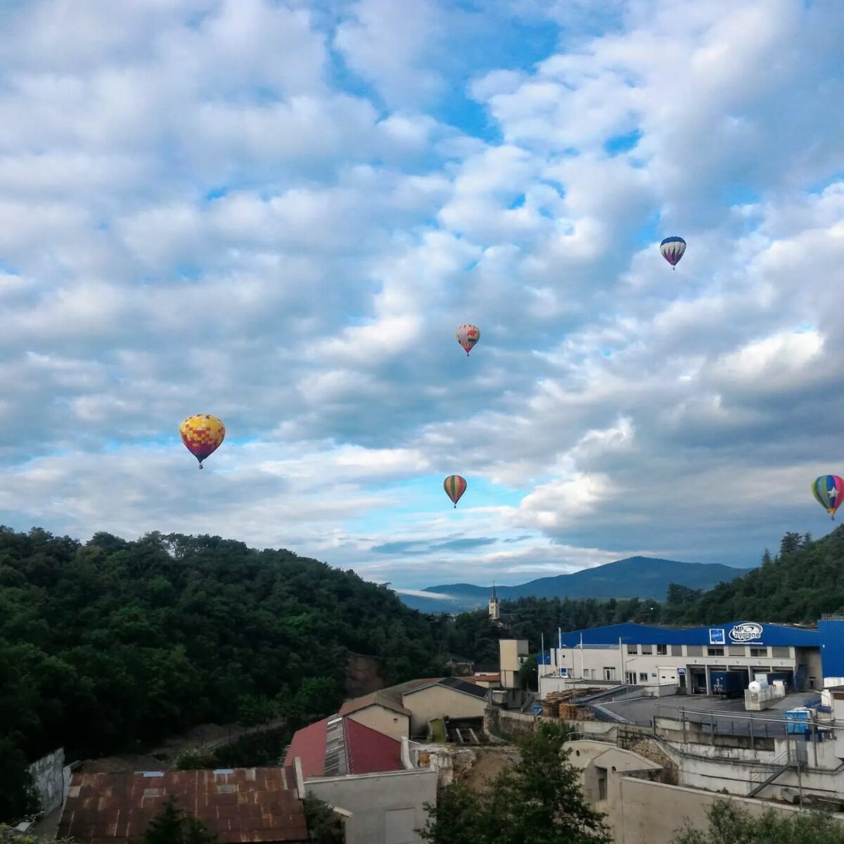 Un essaim de 5 montgolfières dans le ciel au dessus de la vallée de la Deûme avec l'usine MP hygiène au premier plan, l'église de Vidalon et au fond le Suc de Vent et les monts du Vivarais