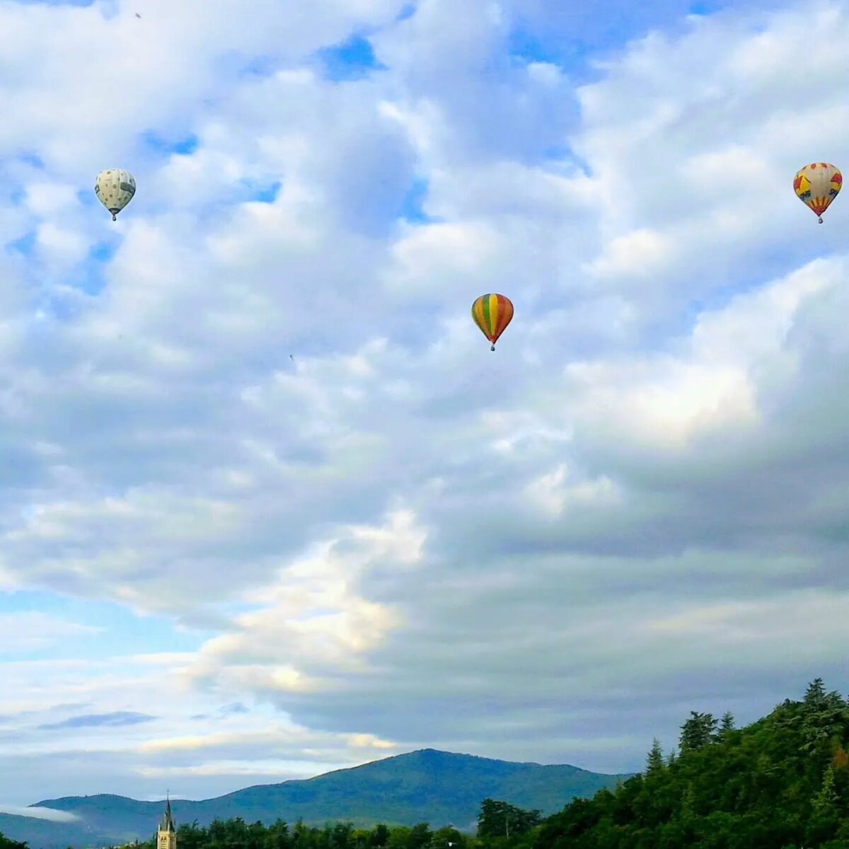 Dans un ciel bleu et nuageux, 3 montgolfières s'envolent au dessus de la vallée de la Deûme et l'église de Vidalon à Davézieux en Ardèche