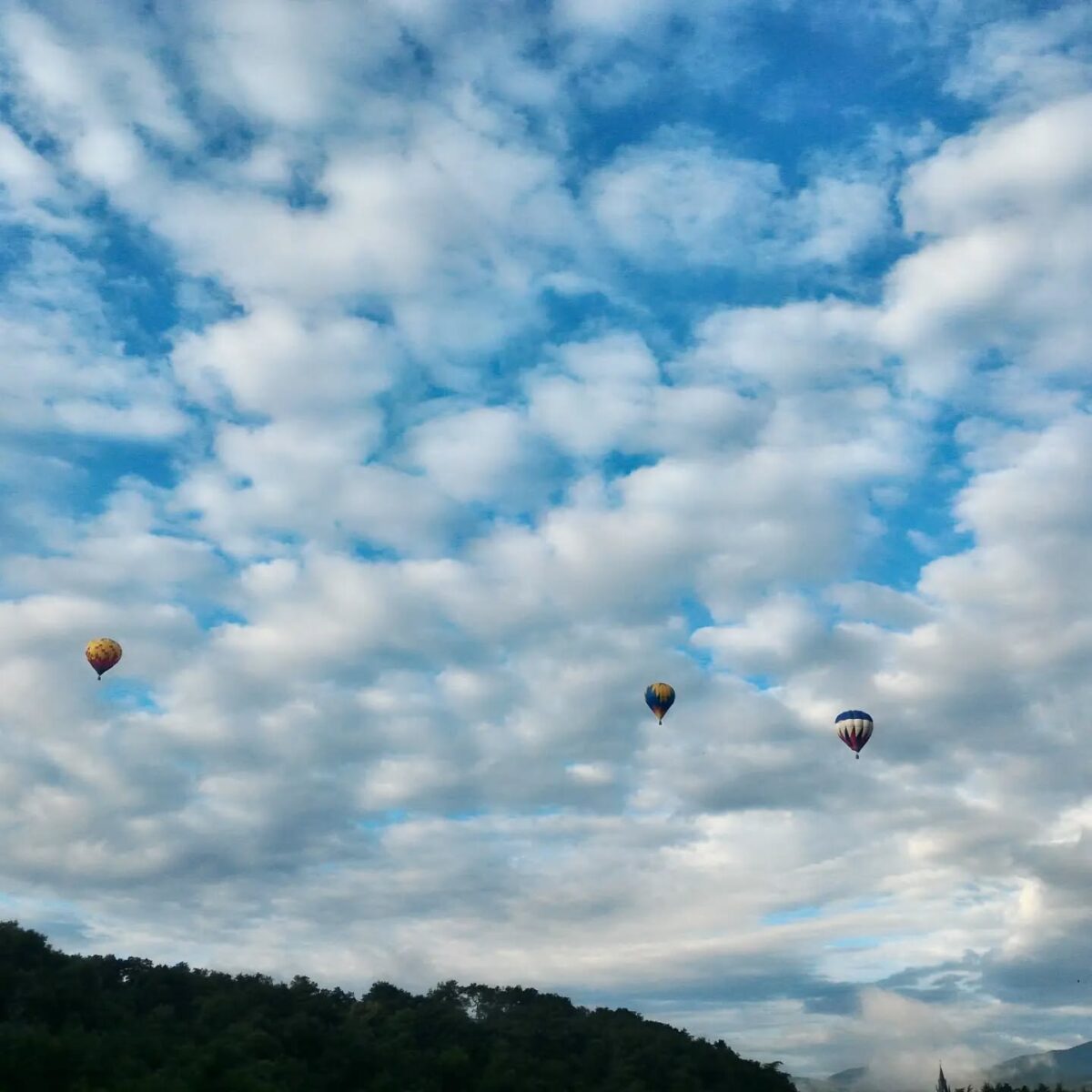 Dans un ciel bleu et parsemés de nuages, 3 montgolfières s'envolent au dessus de la vallée de la Deûme où l'on devine la pointe de l'église de Vidalon à Davézieux en Ardèche