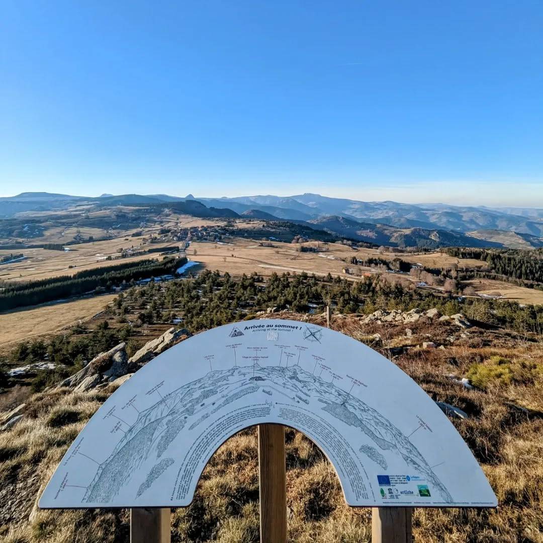 Depuis le sommet du Montivernoux vers l'ouest, vue sur les sucs, le Gerbier de Jonc et le massif du Mézenc avec le village de Lachamp-Raphaël et quelques traces de neige à l'ombre des allées de sapins